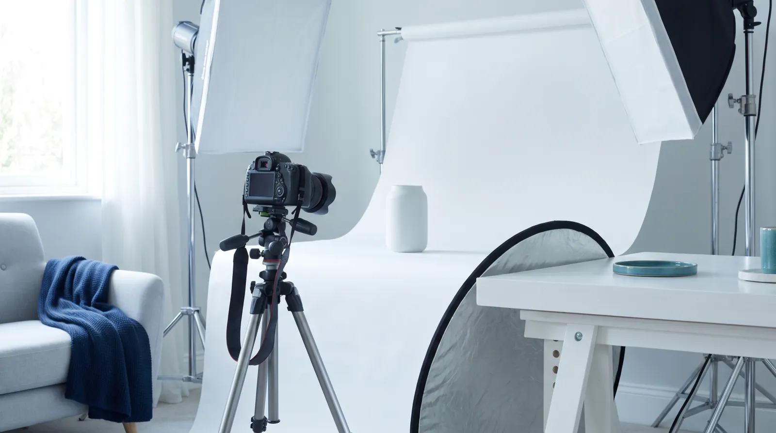 Małe domowe studio: aparat na statywie skierowany na białe tło z matowym białym wazonem, dwie lampy softbox na statywach i blenda oparta o stół, w jasnym świetle.